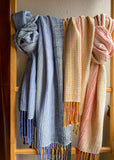 Two woven scarves in soft pastel colors hang from a wooden ladder: a dusty blue scarf on the left, a cream and orange scarf in the middle, and a peach and pink scarf on the right.
